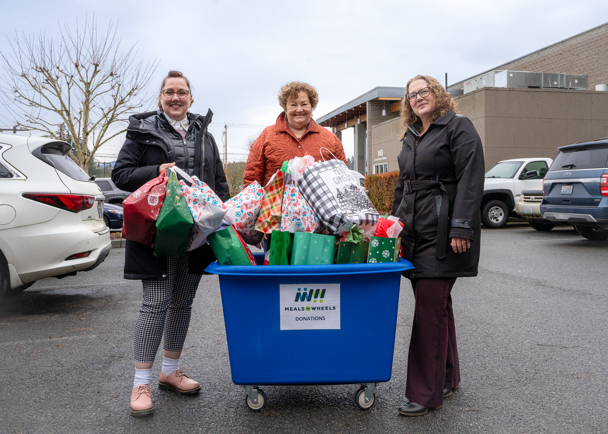 Tres mujeres se encuentran de pie detrás de un carrito de Meals on Wheels con la marca azul, lleno de bolsas de regalo navideñas.