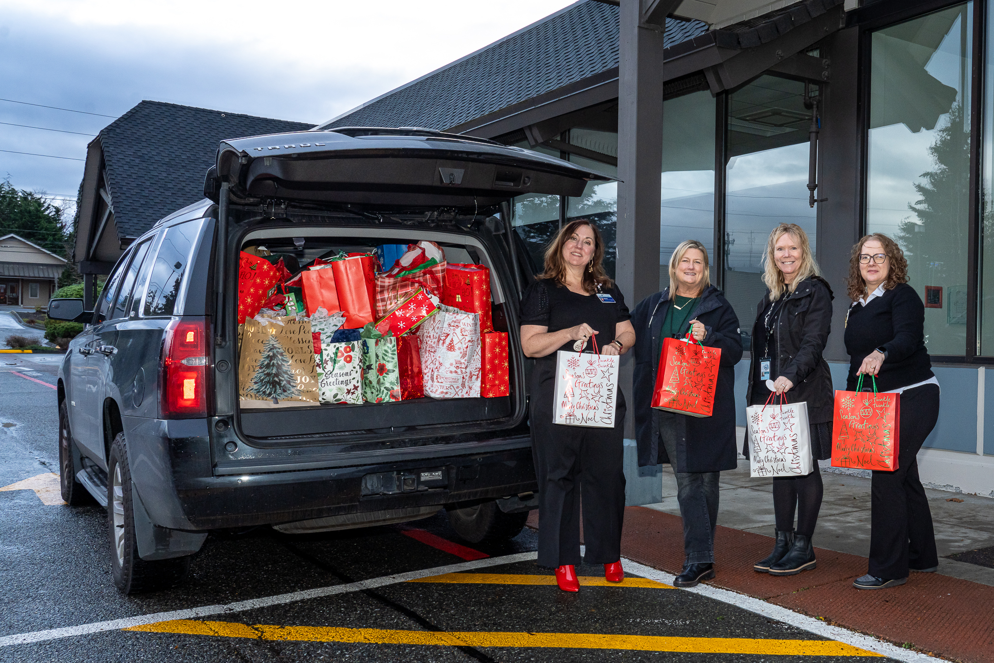 Cuatro mujeres se encuentran de pie a la derecha de una SUV sosteniendo bolsas de regalo rojas y blancas
