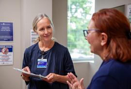 Woman doctor holds paperwork while she listens to woman patient talking in exam room.