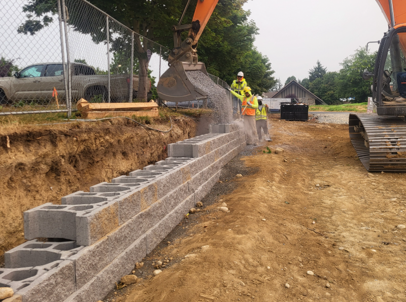 Construction crews install a concrete block retaining wall along an excavated site while an excavator pours gravel behind the wall.