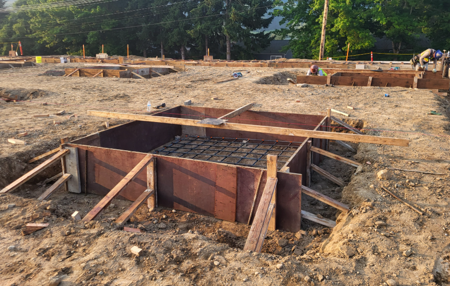 Concrete foundation forms and reinforcing steel are in place across a large construction site as crews prepare for concrete pours. Excavated footings, wooden formwork, and gravel fill are visible throughout the future building footprint.