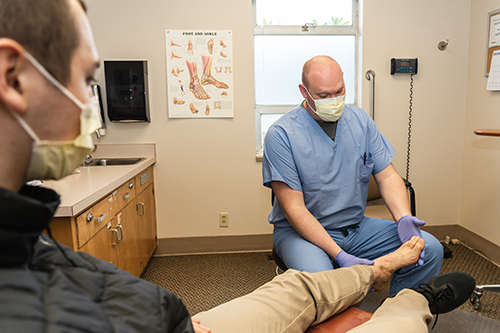 Doctor in blue scrubs examines male patient's foot. 