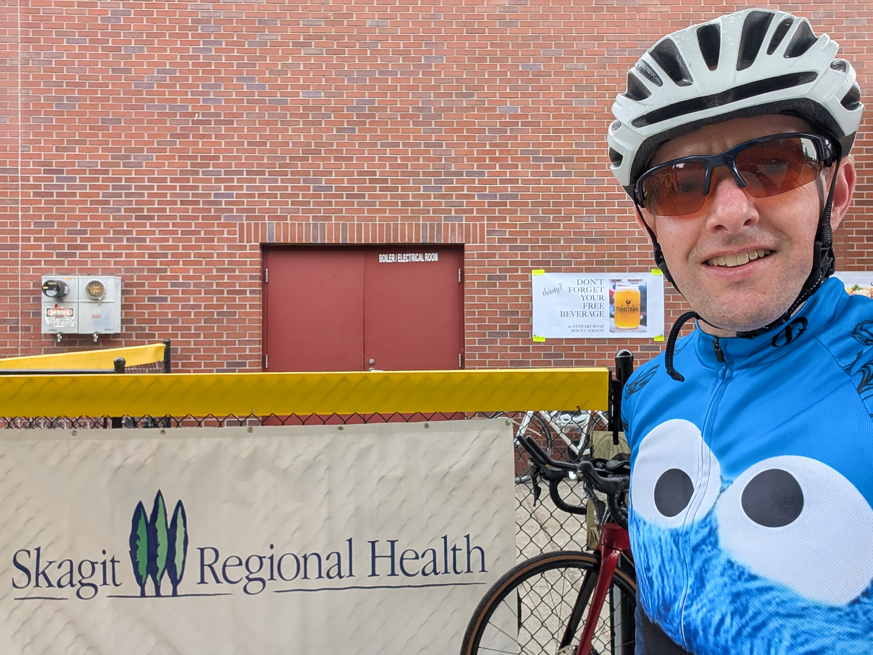 Man in helmet and sunglasses takes photo in front of white Skagit Regional Health banner.