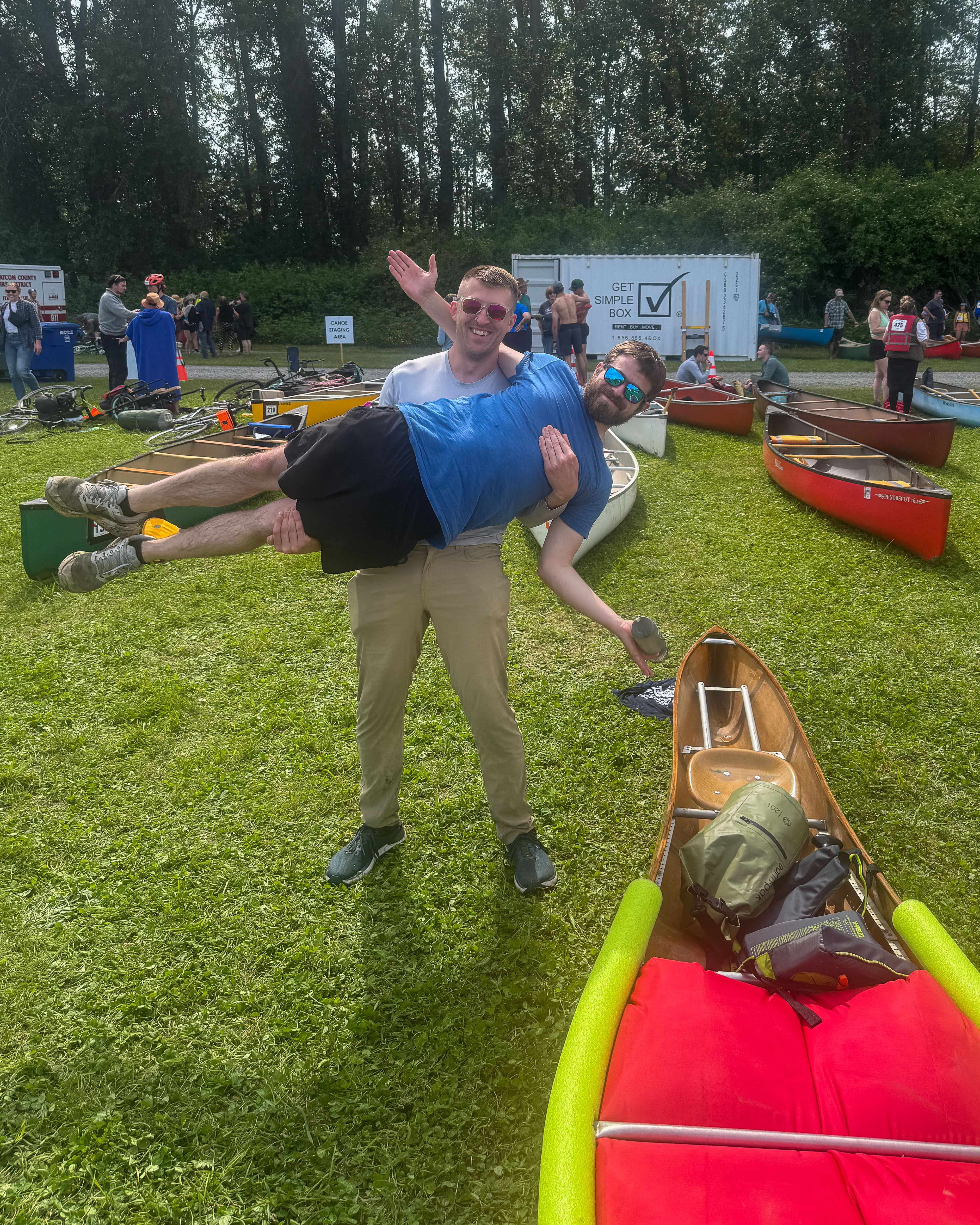 One man stands and holds up another man sideways while standing next to a canoe.