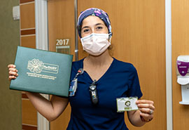 Enfermera con bata azul posa para la foto sosteniendo el premio DAISY Award verde y el pin DAISY.