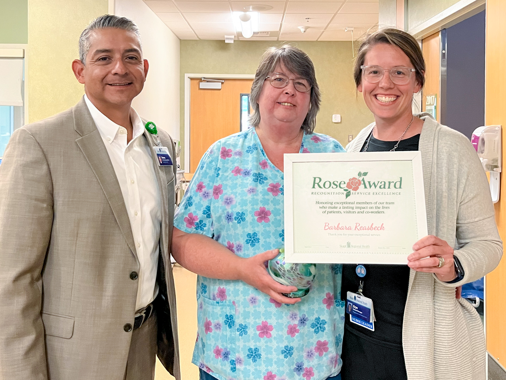 One man, two women pose for photo. Women are holding Rose Award certificate frame