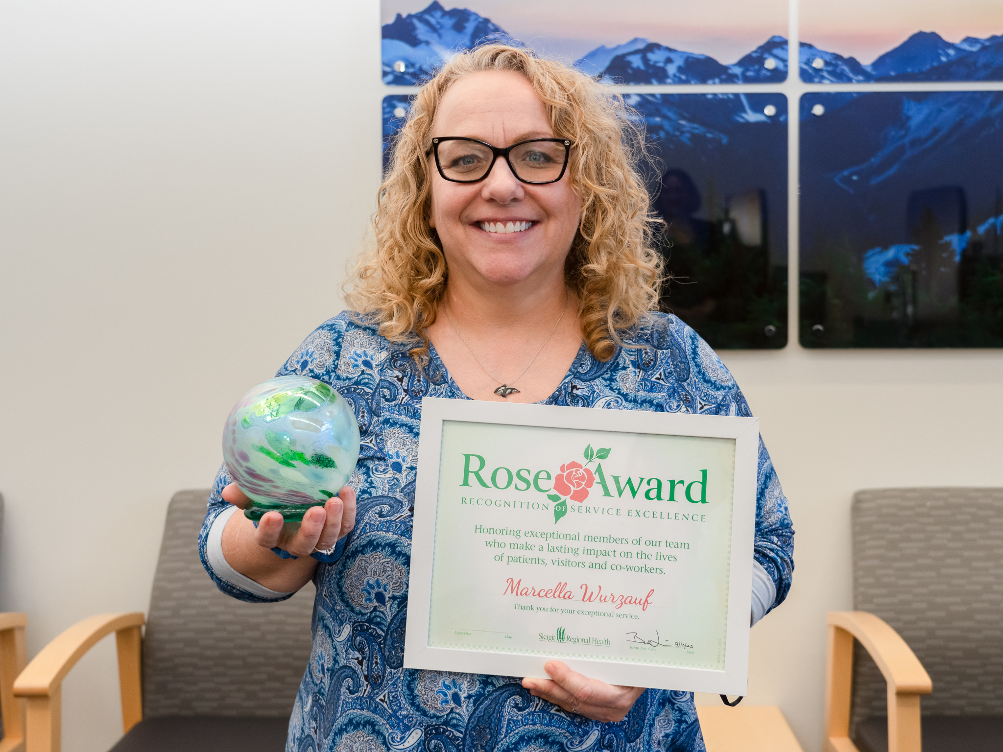 Woman in blue blouse holds Rose Award certificate and blown glass sphere. 