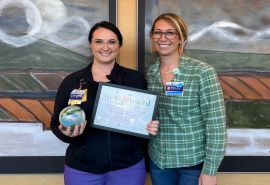 Two women pose in front of two framed pieces of art. Woman on the left holds Rose Award and certificate.