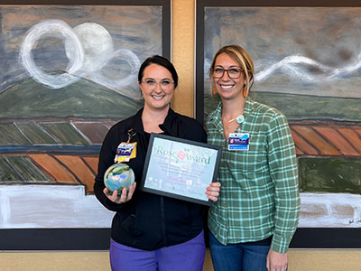 Two women pose in front of two framed art pieces. Woman on the left holds Rose Award and certificate.