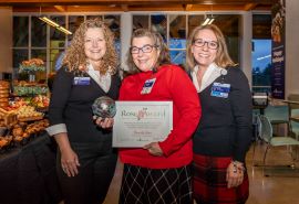 Tres mujeres posan para la foto. Beverly está en el medio, viste un suéter rojo y sostiene su certificado y el premio Rose Award.
