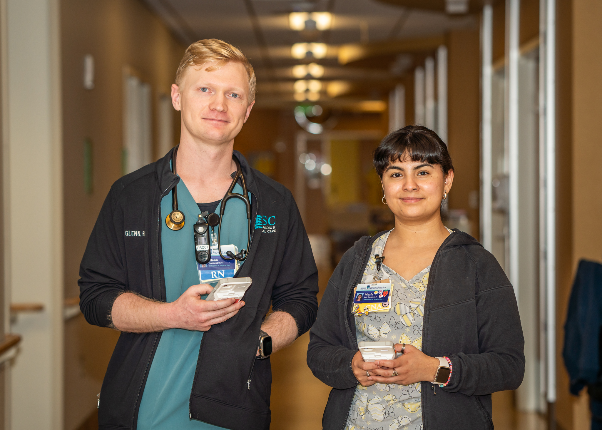 Male nurse and female nursing assistant stand side by side smiling and holding iPhones.