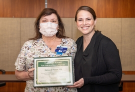 Two women pose together, one holding the DAISY Award certificate and wearing a mask on her face.