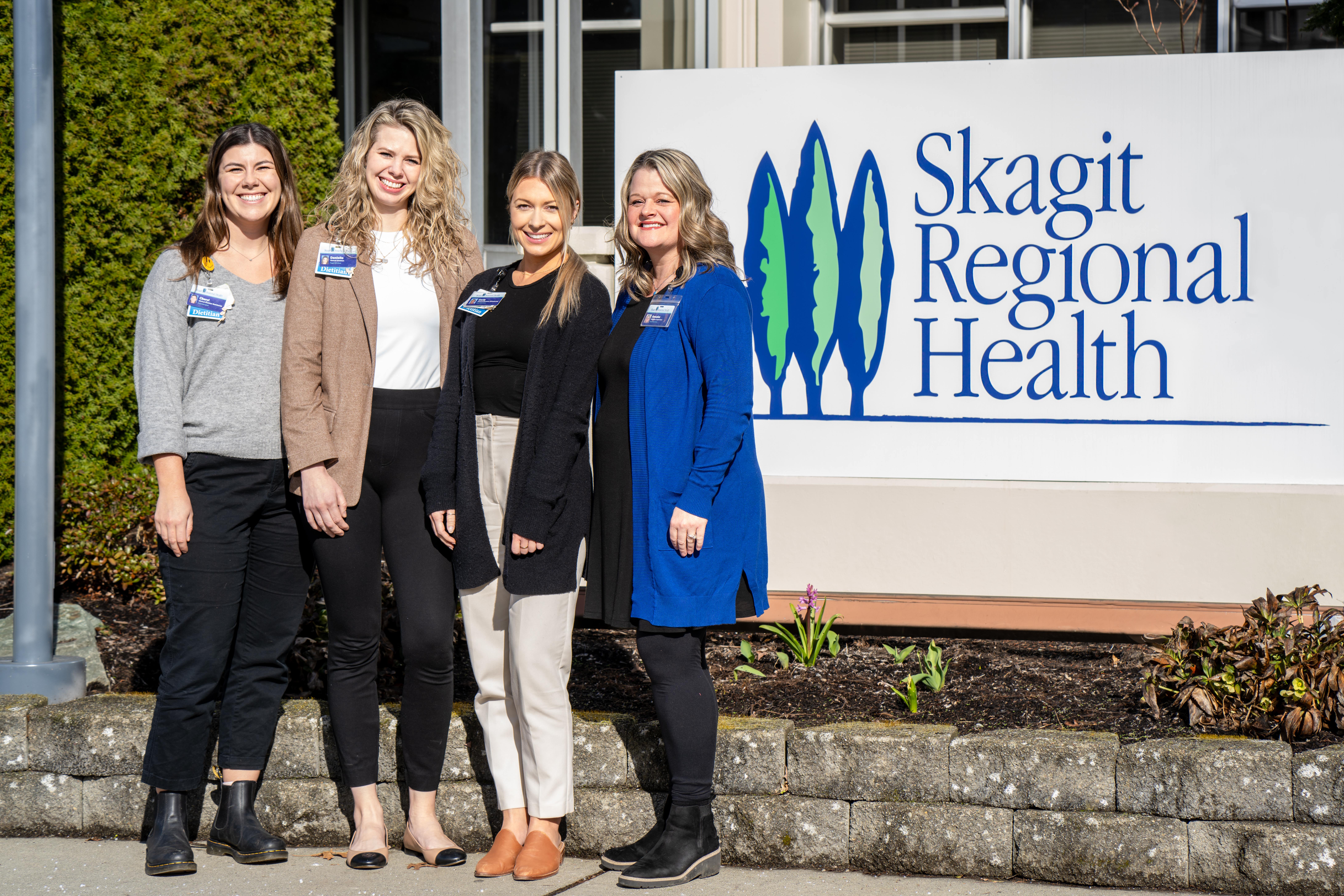 Cuatro mujeres posan juntas a la izquierda del cartel de Skagit Regional Health en exteriores.