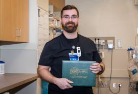 Un enfermero con bata negra sostiene el certificado del premio DAISY Award y posa en la sala de urgencias.