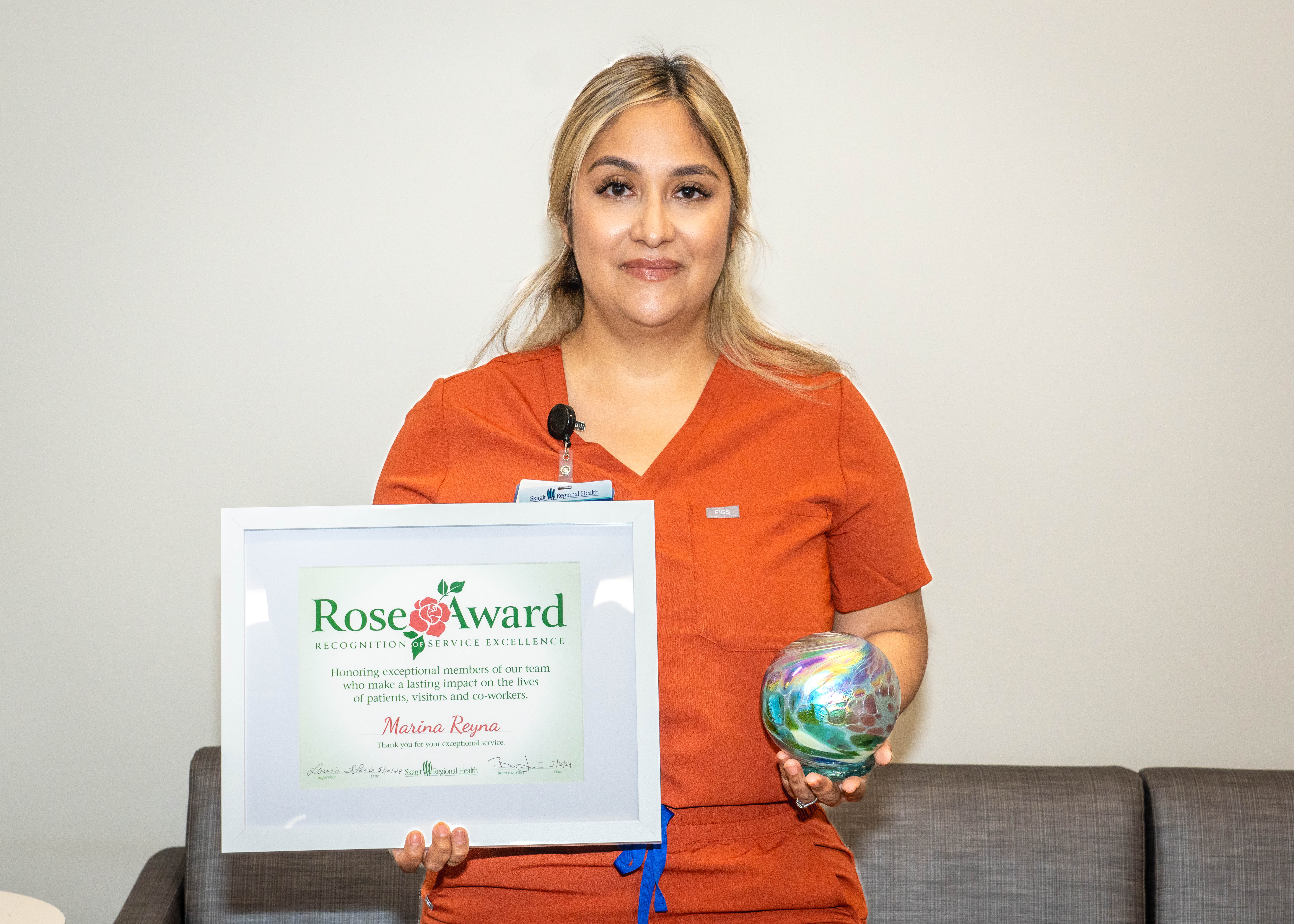 Una mujer vestida con una bata naranja sostiene el premio Rose Award y un certificado.