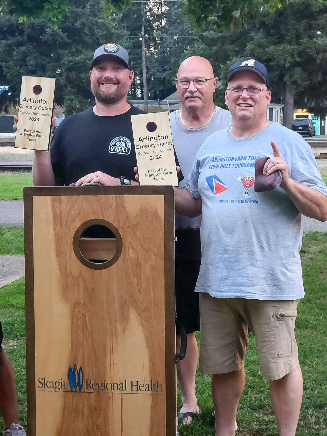 Three men stand behind cornhole board holding up awards at cornhole tournament.