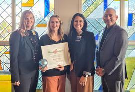 Four people standing in front of a colorful stained-glass window, with one woman in the middle holding a framed Rose Award certificate and an iridescent glass award.