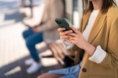Unrecognizable modern young woman holding mobile phone while sitting at bus station and waiting for bus, using smart phone