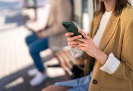 Mujer joven moderna irreconocible sosteniendo un teléfono móvil mientras está sentada en la estación de autobuses esperando el autobús, usando un teléfono inteligente.