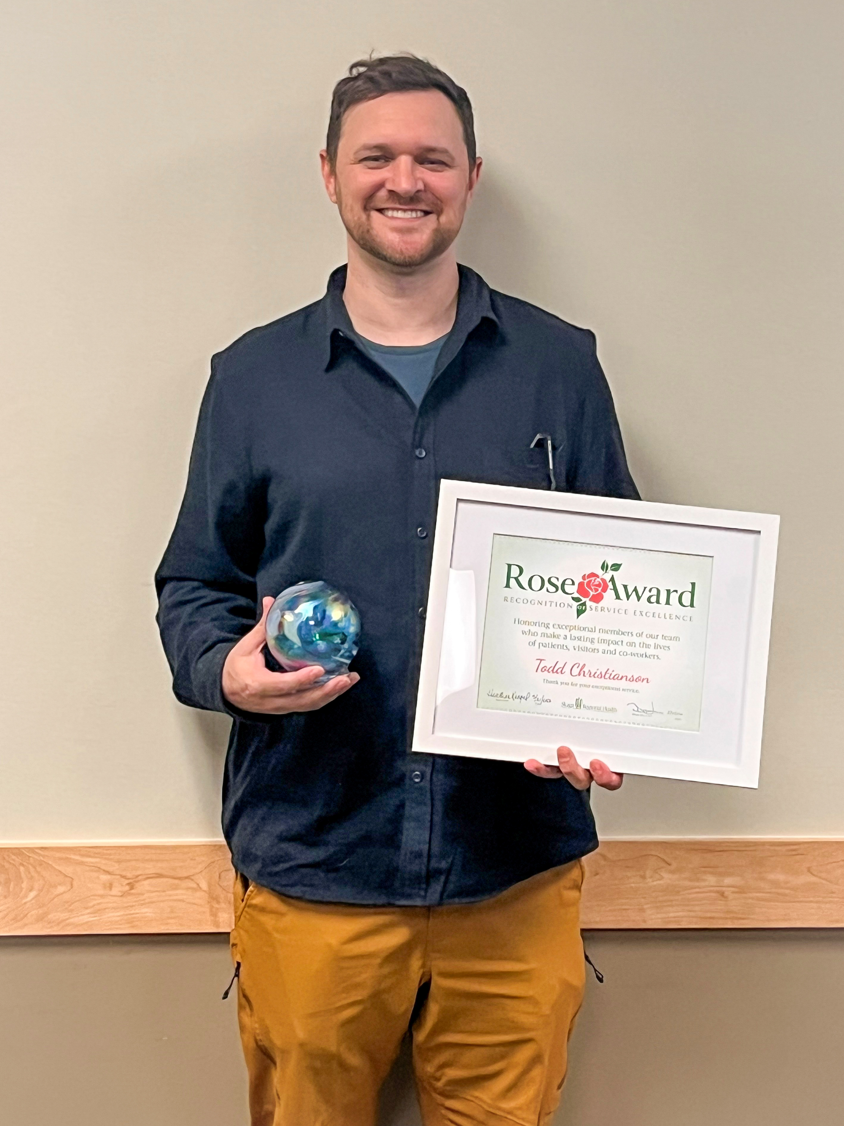 April Rose Award - T. Christianson-2 Man dressed in blue button up and khaki pants stands against a wall holding a Rose Award certificate and blown-glass globe.