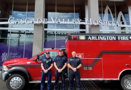 Tres bomberos del Departamento de Bomberos de Arlington posan frente a una ambulancia estacionada frente a la entrada del Cascade Valley Hospital.