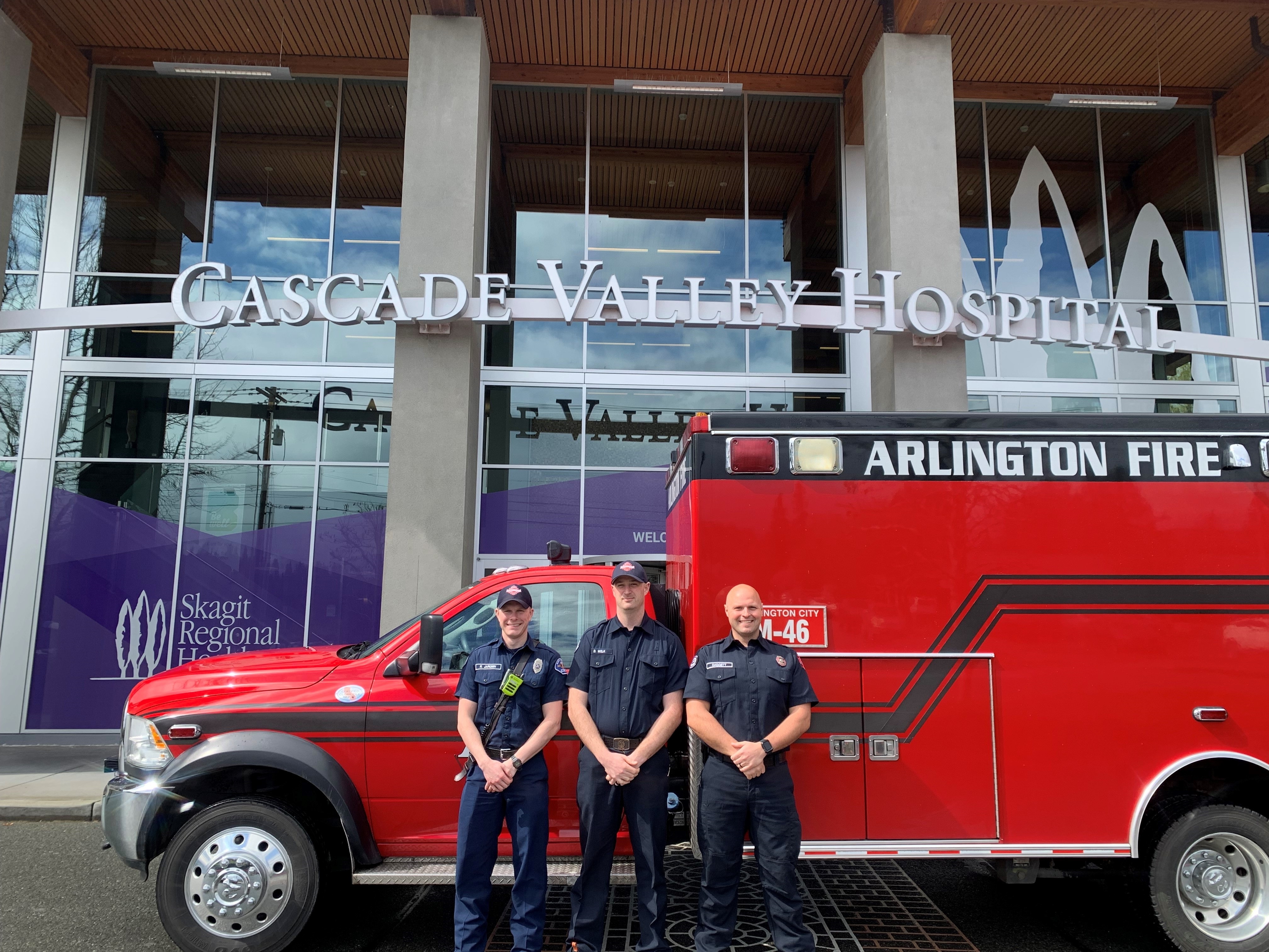Three Arlington Fire Department firefighters pose in front of ambulance parked in front of Cascade Valley Hospital entrance. 