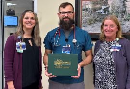Enfermero sostiene su certificado del premio DAISY Award. Dos mujeres junto a él, una de cada lado.