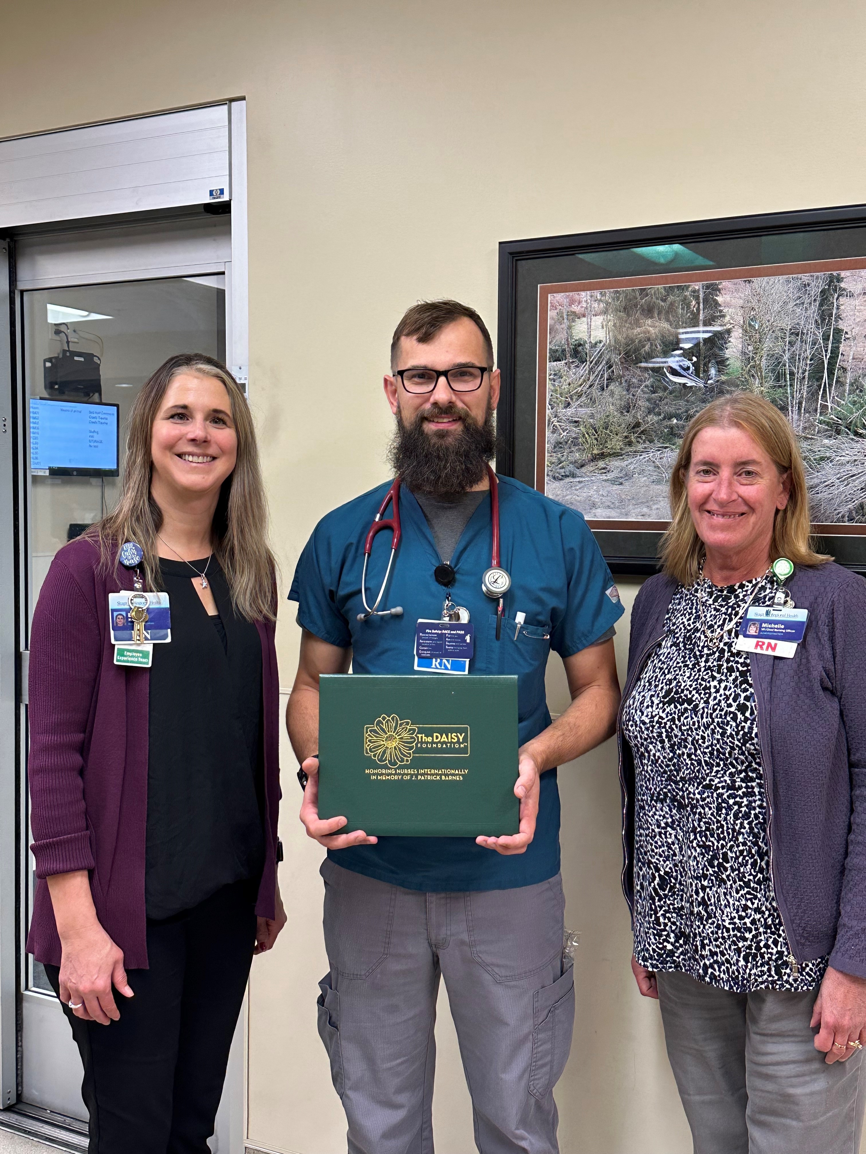 Male nurse holds DAISY Award certificate. Two women next to him, one on each side.