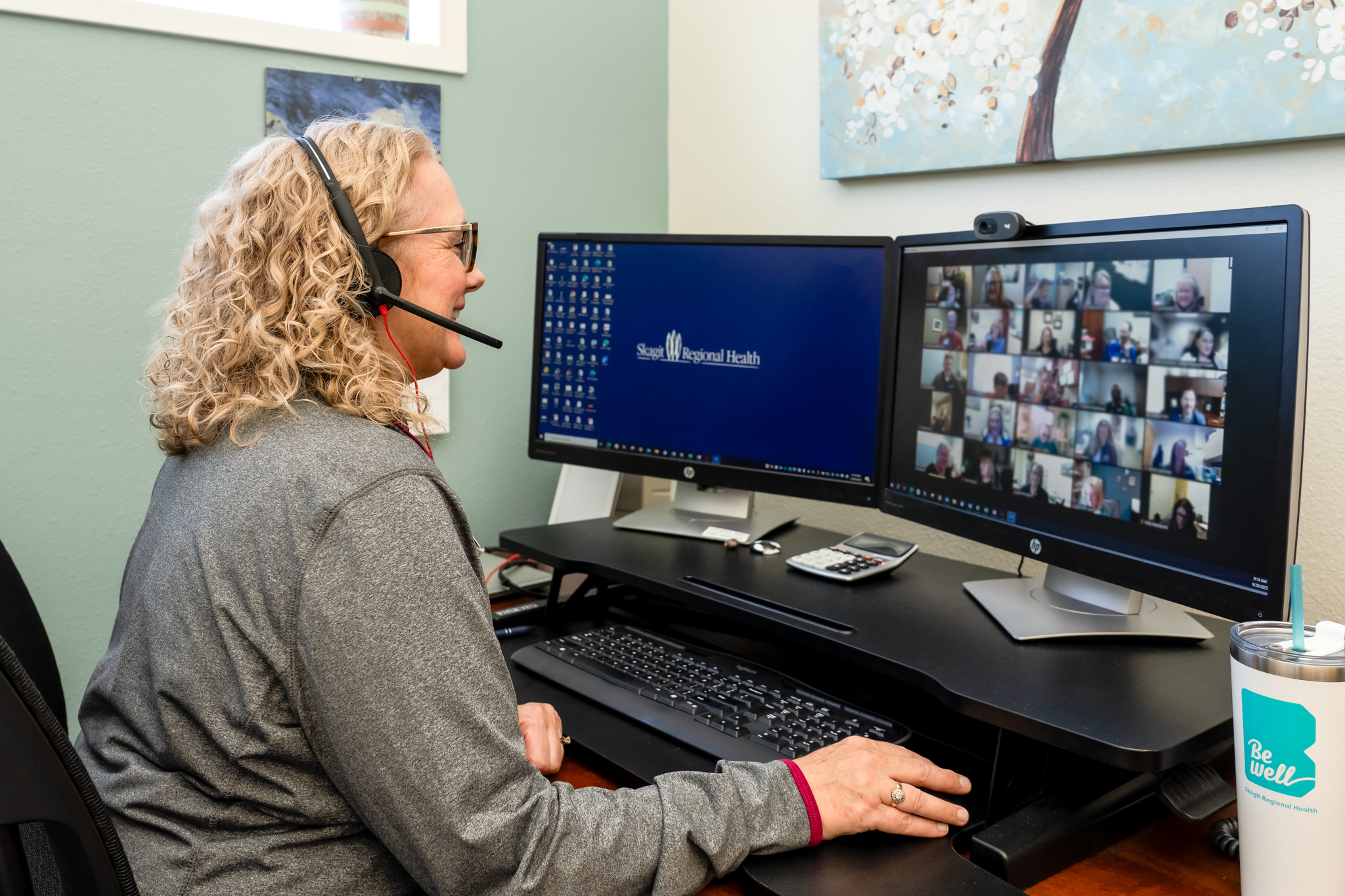 Woman sits at desk smiling at computer screen, screen shows ongoing Zoom meeting. 