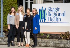 Cuatro mujeres posan a la izquierda del cartel de Skagit Regional Health en exteriores.