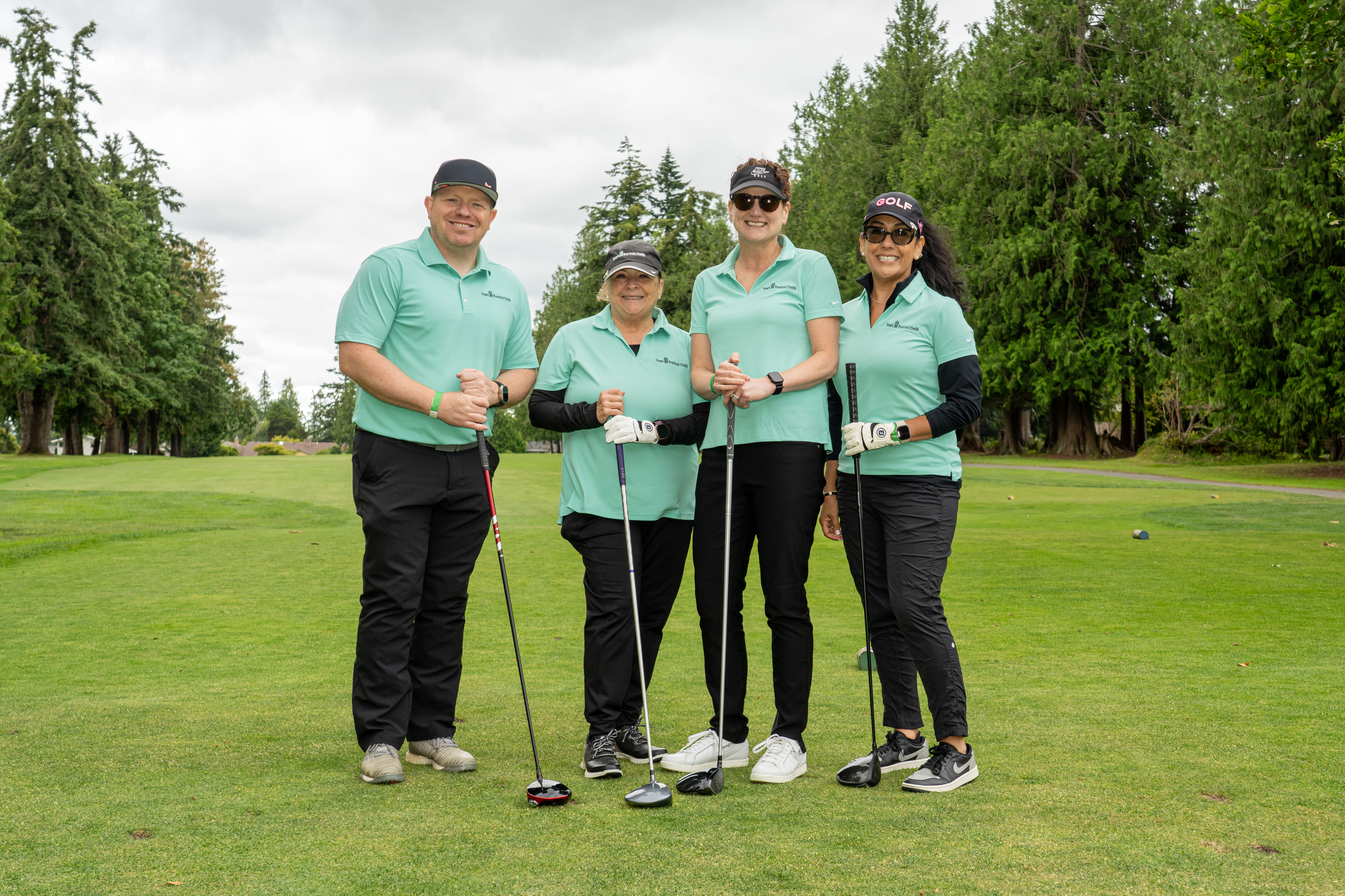 DSC00342 One man and three women holding golf clubs stand on golf course smiling at camera.