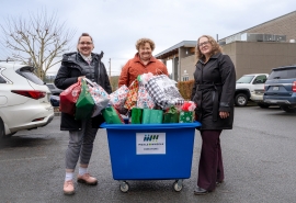 Tres mujeres de pie frente a un carrito de Meals on Wheels con el logo azul, lleno de bolsas de regalo.