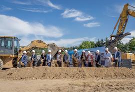 A group of approximately 14 people wearing hard hats stand in a line at a construction site, each holding a shovel in a ceremonial groundbreaking event.