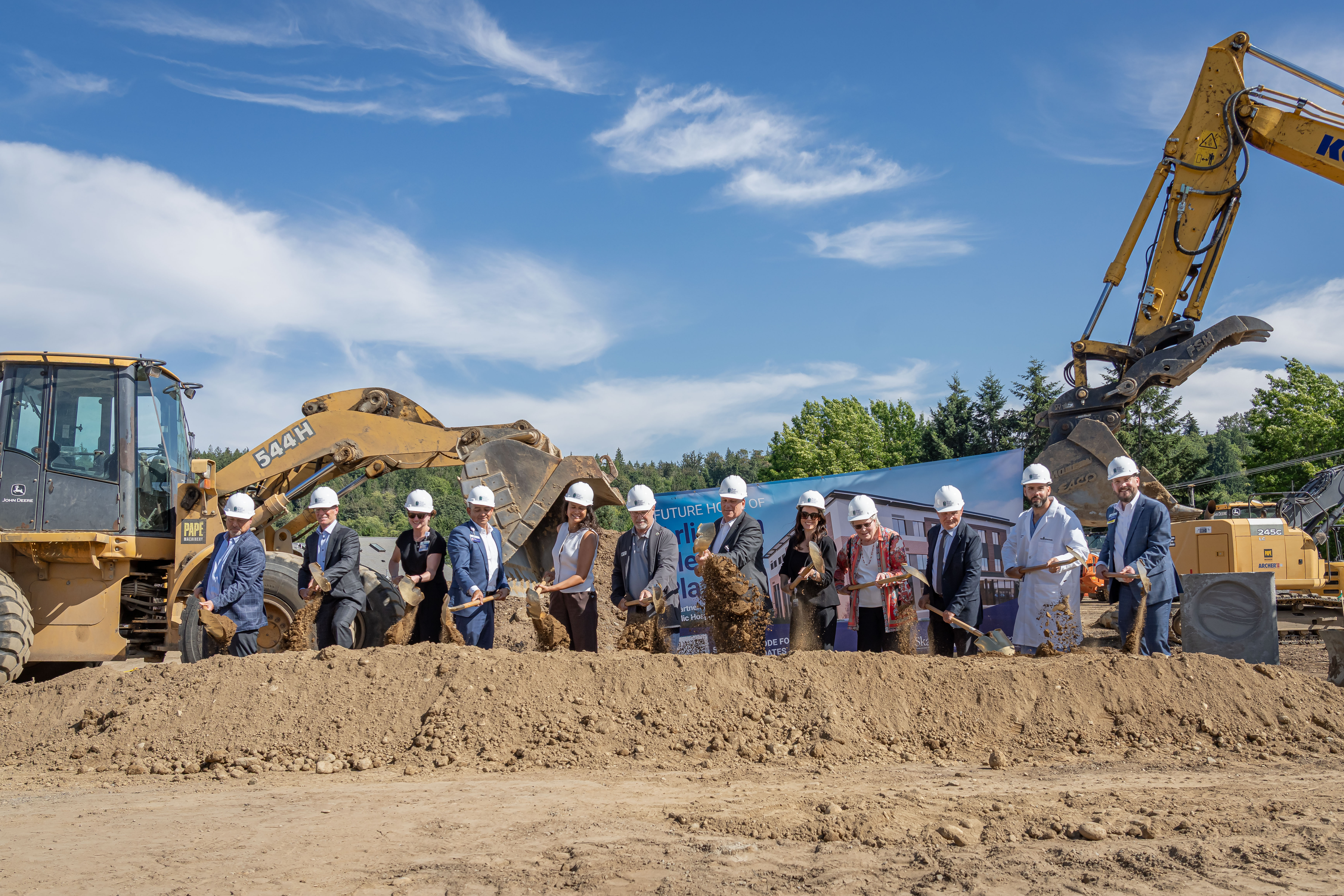 A group of approximately 14 people wearing hard hats stand in a line at a construction site, each holding a shovel in a ceremonial groundbreaking event.