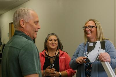 Three people stand in a hallway having a conversation. One person in a green shirt faces two others, one wearing a pink jacket and one holding a screen.