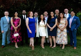 Eleven people in formal attire pose for a group photo on a lawn with trees and lush greenery in the background.