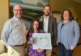 Two men and two women pose for camera, with the woman in the middle holding a framed certificate and blown glass globe.