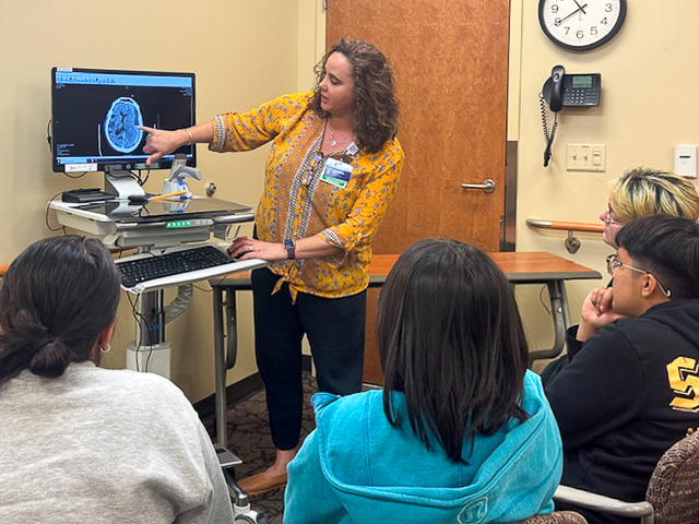 Woman in yellow blouse shows CT scan on computer screen to group of four students.