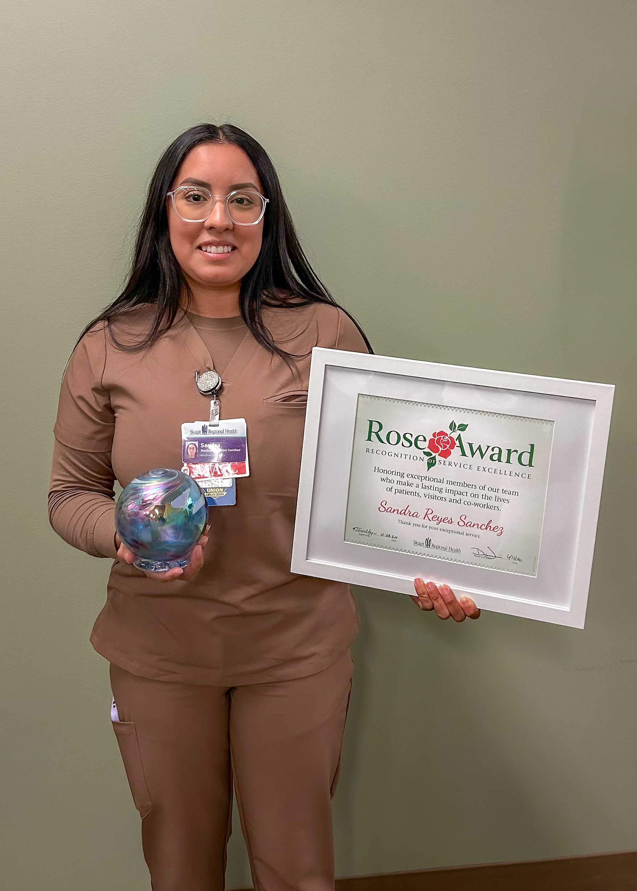 IMG_4320 Woman in brown scrub set stands holding framed Rose Award certificate and blown glass globe art.