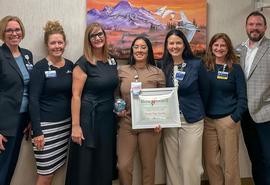 Eight award recipients and presenters pose with a framed certificate and glass award at a recognition event.