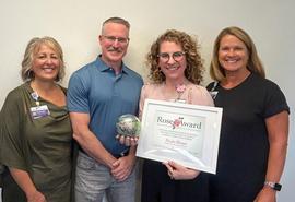 Cuatro personas están de pie en un espacio interior, frente a una pared de color claro. La persona del centro sostiene un certificado del premio Rose Award enmarcado, mientras que otra persona sostiene un premio decorativo de cristal.