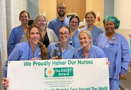 Un grupo de enfermeras con ambos azules posan juntas sonriendo a la cámara, mientras dos enfermeras en la primera fila sostienen un afiche blanco.