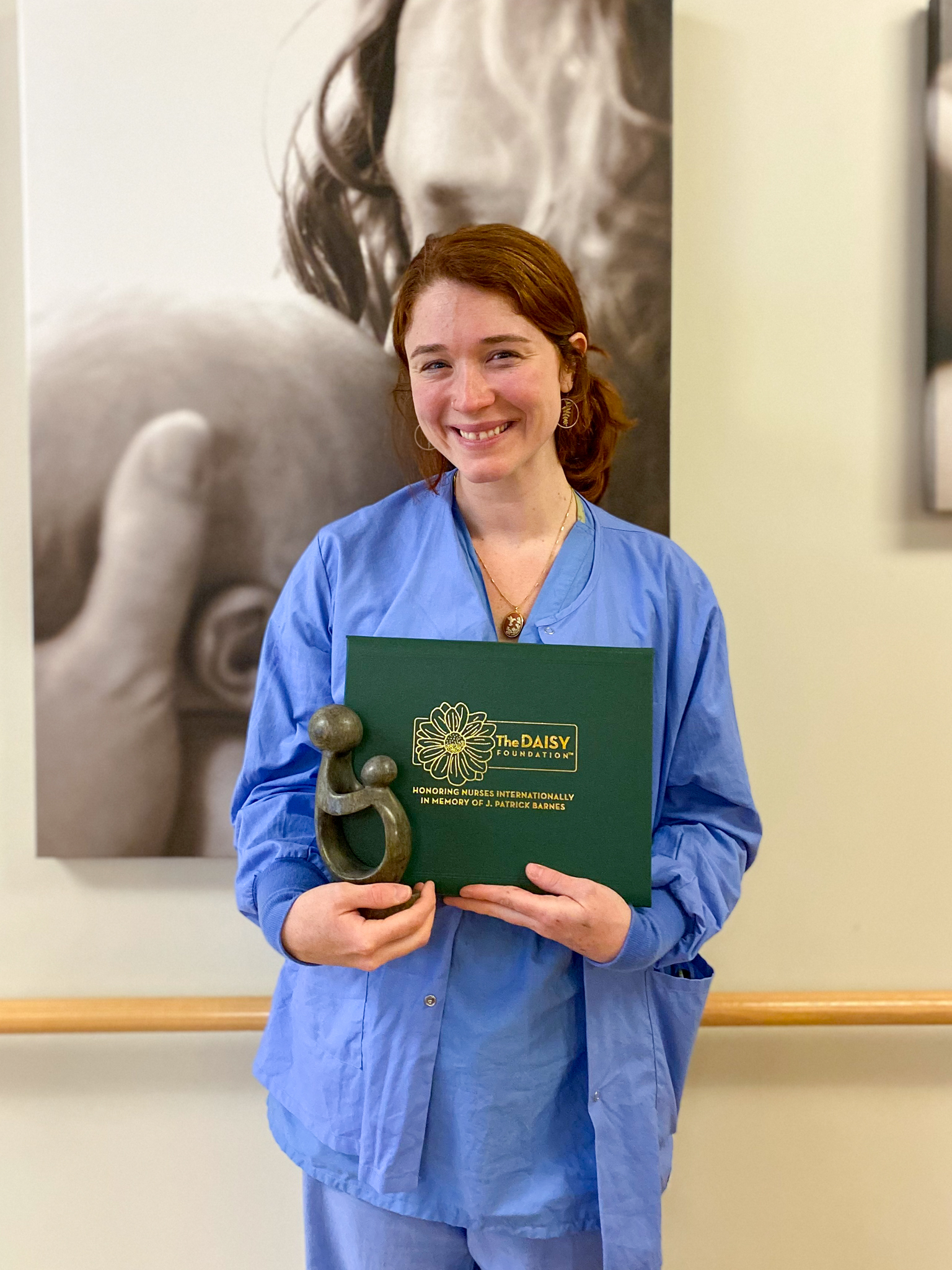 IMG_7430 Female nurse in blue scrubs holds DAISY Award certificate and Healer's Touch statue.