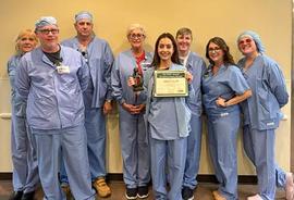 Eight healthcare workers in light blue scrubs standing indoors against a beige wall, with one female nurse holding a DAISY Award certificate in one hand and a DAISY Award sculpture in the other.