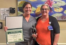 Two women stand in front of blue artwork holding certificate and Healer's Touch statue.