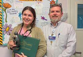 Two people stand side by side indoors in front of a decorated bulletin board. The person on the left is holding a green certificate folder and a small award statue.