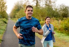 Man and woman run on outdoor path, man leading a few steps ahead of woman.