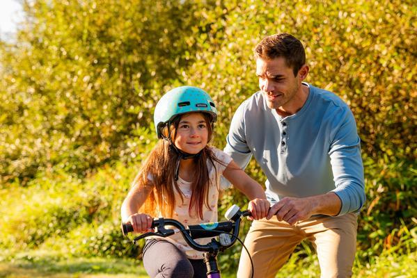 L1440080 Un adulto ayuda a una niña pequeña a aprender a andar en bicicleta en un sendero soleado. La niña lleva un casco azul claro y al fondo se ven árboles y vegetación.
