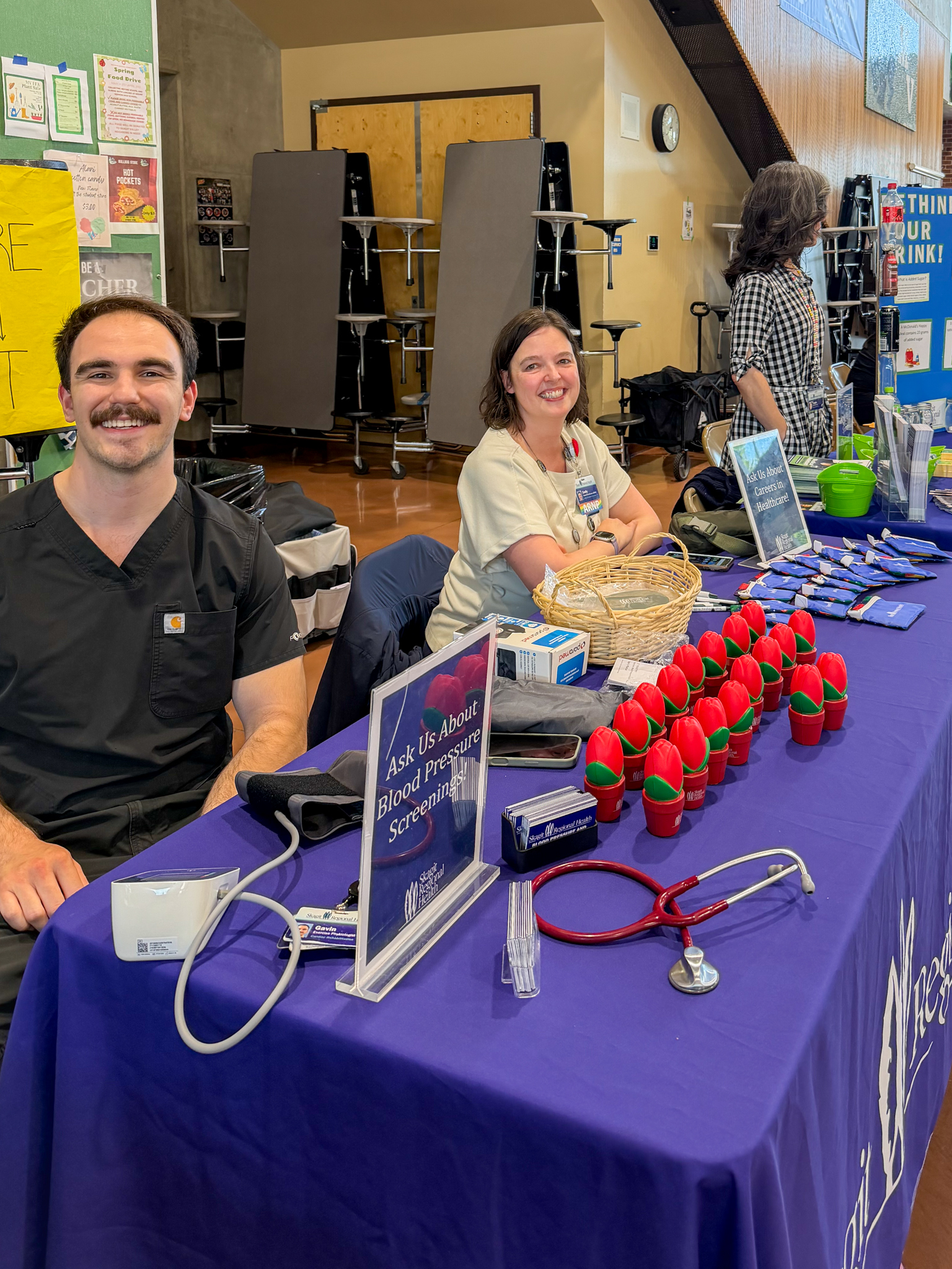 Two people sit behind a table at a community health fair, with a purple tablecloth displaying tulip‑shaped stress balls, pamphlets, pens, and a blood pressure cuff. Other tables and attendees are visible in the background inside a gym‑like setting.