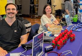 Two people sit behind a table at a community health fair, with a purple tablecloth displaying tulip-shaped stress balls, pamphlets, pens, and a blood pressure cuff. Other tables and attendees are visible in the background inside a gym‑like setting.